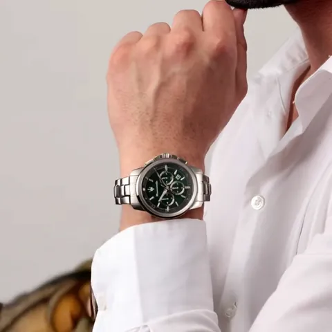 A man's wrist at the cuff of a white shirt, wearing a watch with a steel bracelet and a dark green dial carrying three c