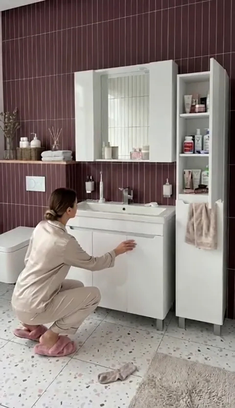 A bathroom in ribbed burgundy tile: a woman in pyjamas crouches to open the lower door of a white vanity. A tall unit wi