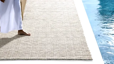 The rug lies right at the edge of a pool: a cream flatweave of coarse texture, water up against the edge. The frame sets