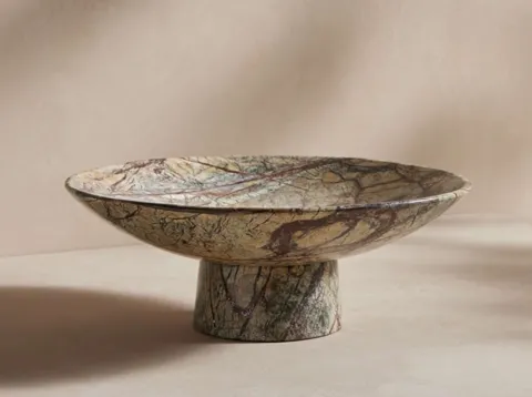 The bowl stands alone on a sand ground: a wide shallow form on a conical foot, pale stone with brown and greenish veinin