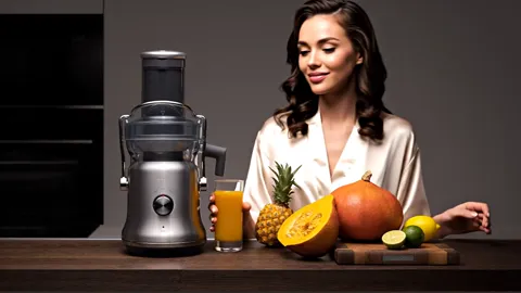 A dark kitchen: a juicer with a steel body and a wide feed chute stands on the worktop beside a pineapple, a pumpkin, li