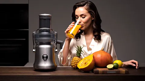 She drinks, the frame closer. The spout and the pulp container at the side are legible — the two details a centrifugal j