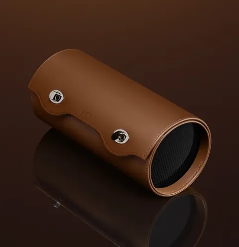 The case lies closed on a dark mirrored surface: brown leather, two steel press studs, a carbon end cap. The form is cyl