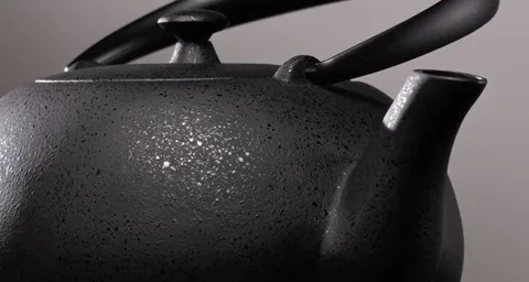 Macro on the side of the pot: black ceramic with a coarse granular relief, a slim spout, a handle arching over the lid. 