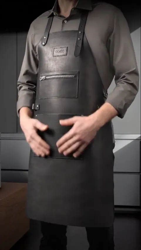 A man in a grey leather apron in a dark kitchen, framed to the chest. A zipped chest pocket and riveted strap fixings ar