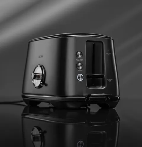 The toaster stands on a mirrored surface: a matte black body, a round chrome browning dial, two slots. The reflection be