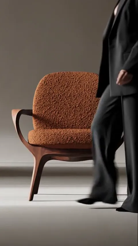 The chair stands alone in a lit studio: a bent wooden frame with arms, seat and back in terracotta bouclé. A woman in a 
