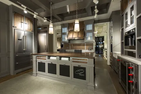 The kitchen entire: grey-green framed fronts, a long island with a wooden worktop, professional appliances in their hous
