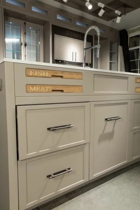 A kitchen in pale grey fronts: an island with a sink, a tall spring-loaded tap, glazed display cabinets above. Every dra