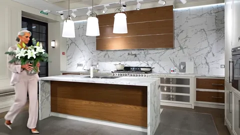 The kitchen entire: a marble splashback and worktop, wooden fronts, an island, pendant lights. A woman in a pale suit en