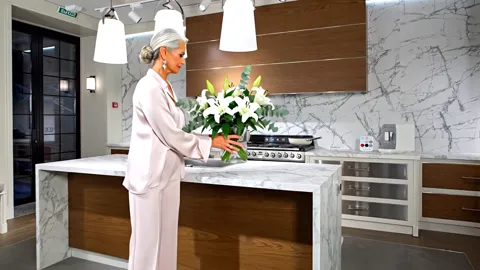 She sets the flowers on the island. Her figure gives the worktop height and the width of the island — things that say no
