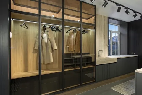 A dressing room: bays with framed glass doors, lit inside, coats and hangers on the rails. A window to the right and a d