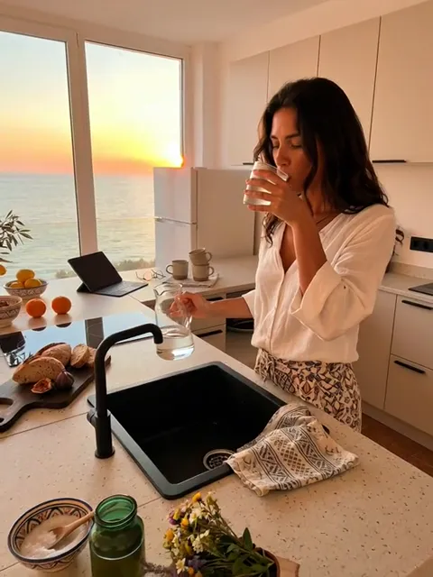 She drinks, the frame closer. The depth of the island reads: a working strip remains behind the sink, not just an edge.