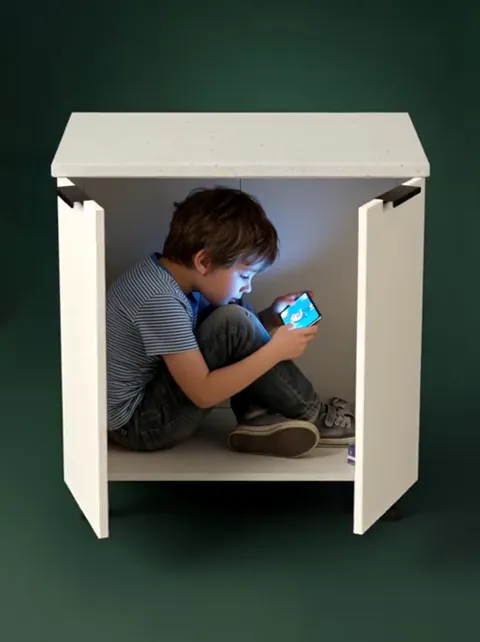The doors open: a boy sits inside with a phone. A child fits into the carcass whole — that is the statement about intern