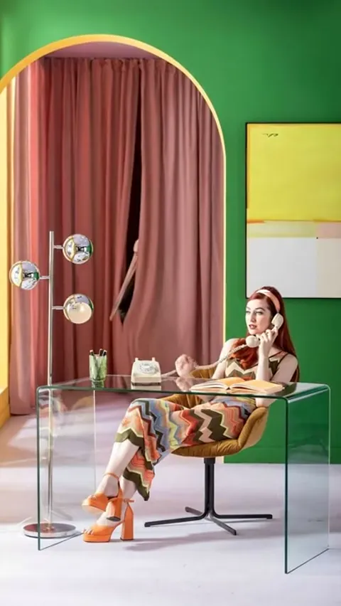 A room with green walls and a pink arch: a woman in a vintage dress sits at a clear glass desk with a telephone handset.