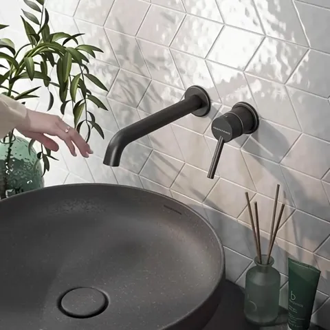 Close: white relief tile in diamonds, a black wall-mounted tap, a dark stone basin. The relief catches the side light — 