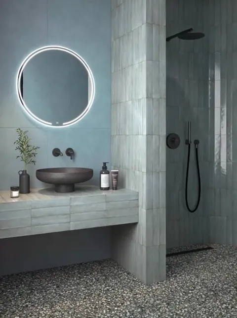 A bathroom in blues and greens: a round backlit mirror, a dark stone bowl on a floating counter, a black wall tap, a sho