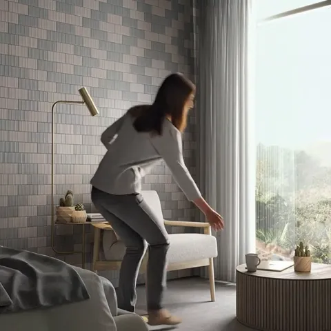 A room with a full-height window and a wall of small brick-format tile in a warm grey. A woman walks to a chair by the w