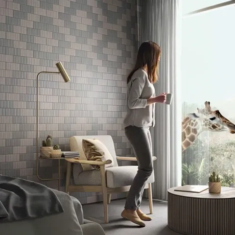 She stands by the window with a cup. A giraffe has appeared outside and stretches its neck toward the room. The tiled wa