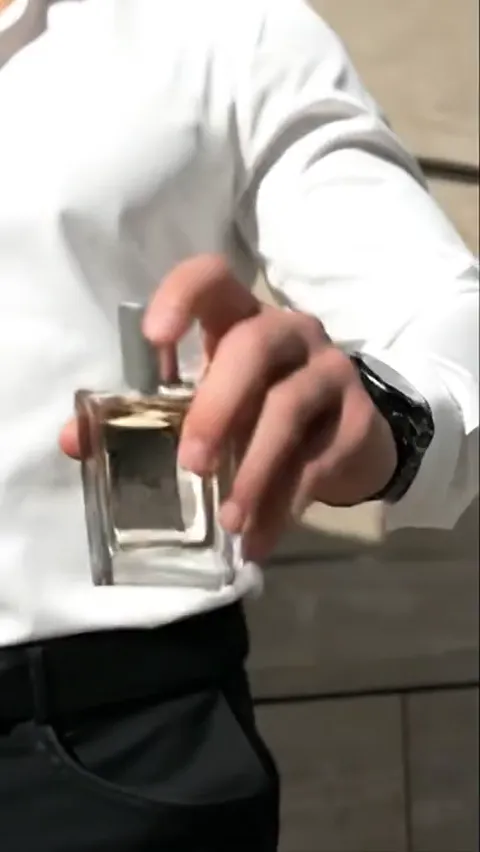 A man in a white shirt holds a bottle out to the camera, framed to the chest. The bottle in focus, his face soft behind 