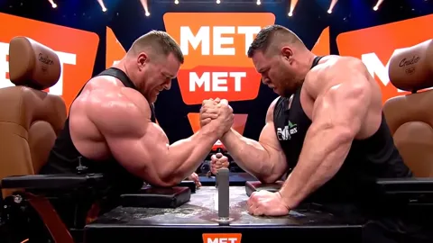 Close: two men arm-wrestling at a table, MET logos on the backdrop, stage lighting. The frame reads as coverage of a spo