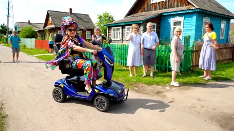 A village street of wooden houses: a blue four-wheeled scooter drives along the dirt road, an elderly woman in bright sc