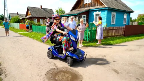 The scooter passes the onlookers. Four wheels give stability on the dirt — the machine does not lean on the uneven surfa