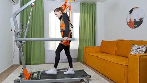 A rehabilitation room: a person in a suspension harness walks on a treadmill, the trainer's orange frame carrying them f