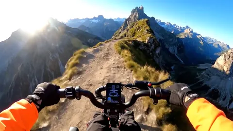 A first-person view: gloved hands on the bars, a narrow ridge trail ahead. A valley below, the sun straight into the len