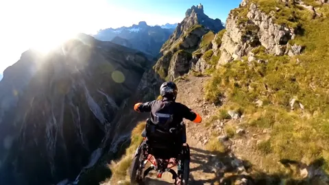 The frame cuts to an external view: a rider on a handcycle attachment coupled to a wheelchair. The front wheel drives, t