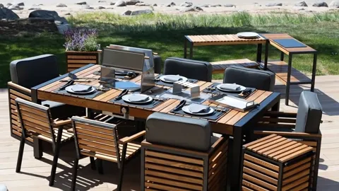 A wooden deck at the water's edge: a long slatted table with dark-cushioned chairs around it, a grill set into the top. 