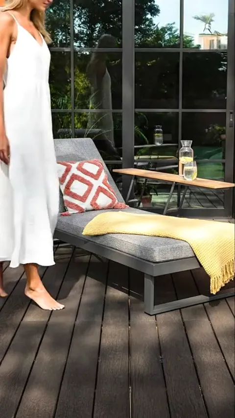 A dark wooden deck by a glazed wall: a grey sun lounger with a mattress and raised headrest, a side table with a carafe.