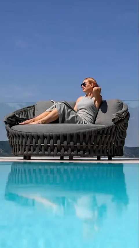 A poolside terrace with mountains beyond: a round woven daybed with a tall curved hood, grey cushions inside. A woman re