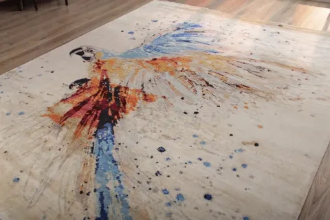 The rug lies in a bright room: a watercolour parrot on a cream field, in spatters of blue, rust and black. The light is 