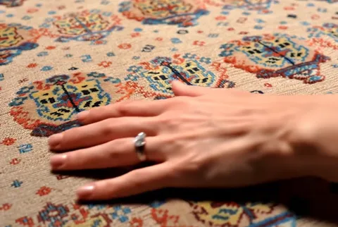 Close: the cream field of a kilim with medallions in blue, yellow and red. A woman's hand enters and settles onto the we