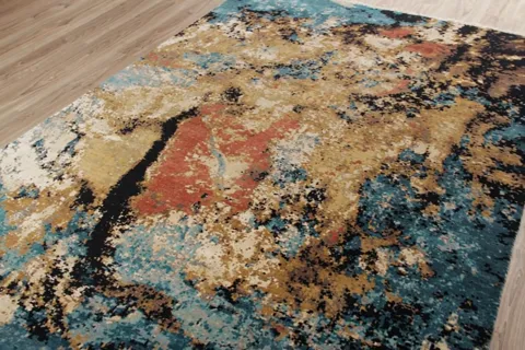 The rug lies on a pale floor: an abstract field of turquoise, ochre, black and terracotta patches, as though spilled acr