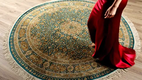 A round rug in front of a fireplace: a turquoise and gold medallion of concentric ornament, fringed at the edge. A woman
