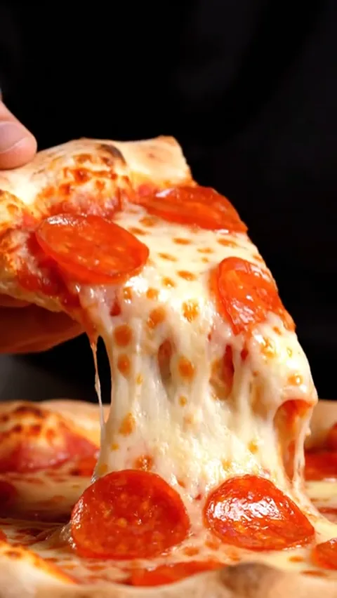 A close shot: a hand lifts a slice of pepperoni pizza, strings of cheese trailing behind. The ground is black; only food