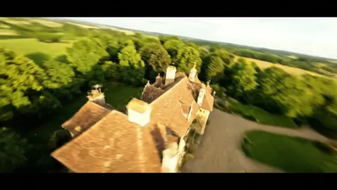 An aerial shot: a stone manor with a tiled roof among green fields and copses. The camera travels over the treetops.