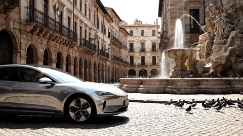 An Italian piazza with a baroque fountain and arcades: a silver electric car crosses the cobbles. The bodywork reflects 