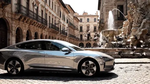 The car in profile, the fountain behind. The body is smooth, with flush handles and closed wheel covers — aerodynamics r