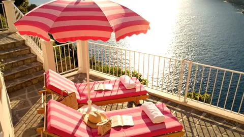 A terrace above the sea: a pink-and-white striped parasol, two loungers with towels, a straw hat. Water and a rocky shor