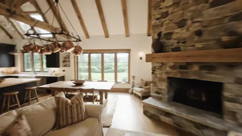 The living room of a country house: a stone fireplace across the wall, timber beams overhead, a pale sofa. The camera mo