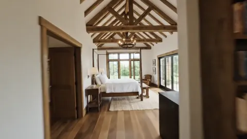 The camera moves into a bedroom: the same beams, a window onto the garden, a bed against the wall. One ceiling material 