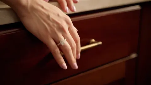 A woman's hand reaches for a drawer handle. A diamond ring on the ring finger, window light sliding across the stone.