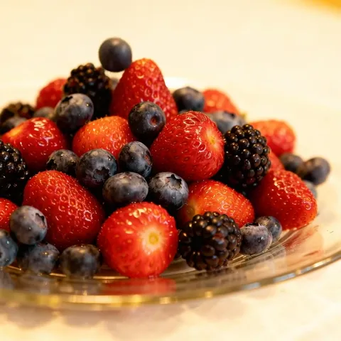 A glass plate of berries, close: strawberries, blueberries and blackberries in a mound. A warm pale ground; nothing but 