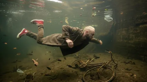 Underwater: an elderly man in a coat and red sneakers hangs horizontally above a silted bed. Fine particles and fish aro