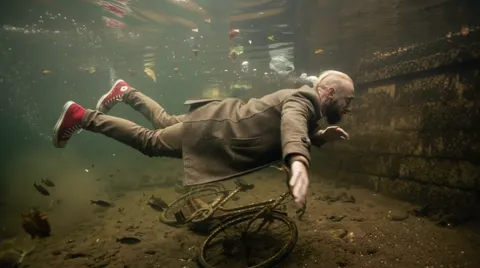 A sunken bicycle appears below. A second object gives the scene its depth and the figure its scale.