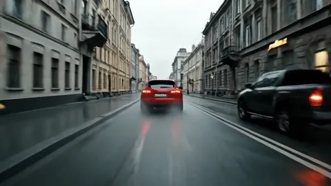 From behind along the street: the car pulls away between the buildings. The width of the roadway against the body is vis