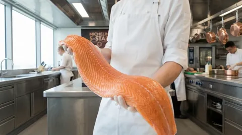 A professional kitchen: a chef in whites holds a salmon fillet across both hands. Colleagues and steel surfaces behind.